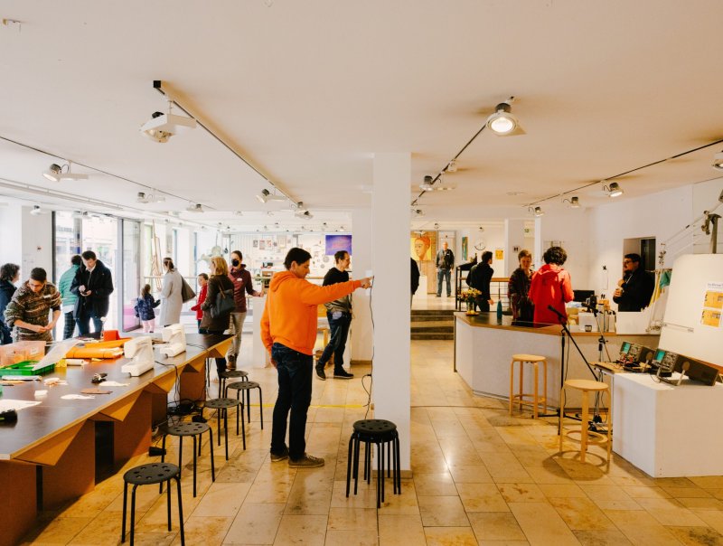 Innenansicht eines Ausstellungs- und Workshopraums mit mehreren Besuchern, die sich um Stühle und Tische gruppieren.