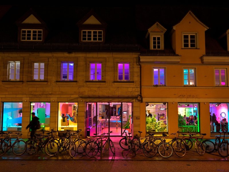 Fassade des ZAM-Gebäudes bei Nacht mit vielen Fahrrädern davor. Innenräume sind sichtbar und bunt beleuchtet.