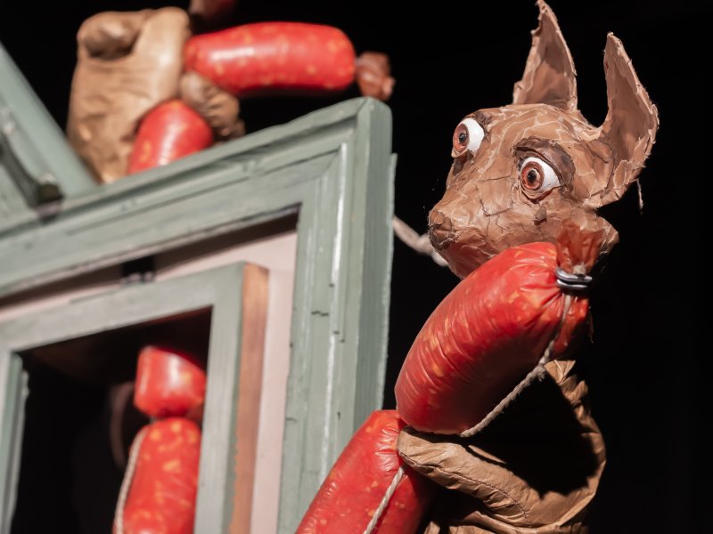 Eine Maus aus Pappmaché, die eine rote Wurstkette hält und hinter einem Fensterrahmen hervorschaut. Im Hintergrund eine weitere nur teilweise zu sehende Puppe, die ebenfalls die Wurstkette umklammert.
