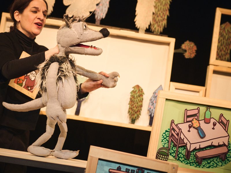 Puppenspielerin führt eine graue Wolf-Puppe auf der Bühne vor. Im Hintergrund sind bemalte Kulissen mit Möbeln und Pflanzen sichtbar.