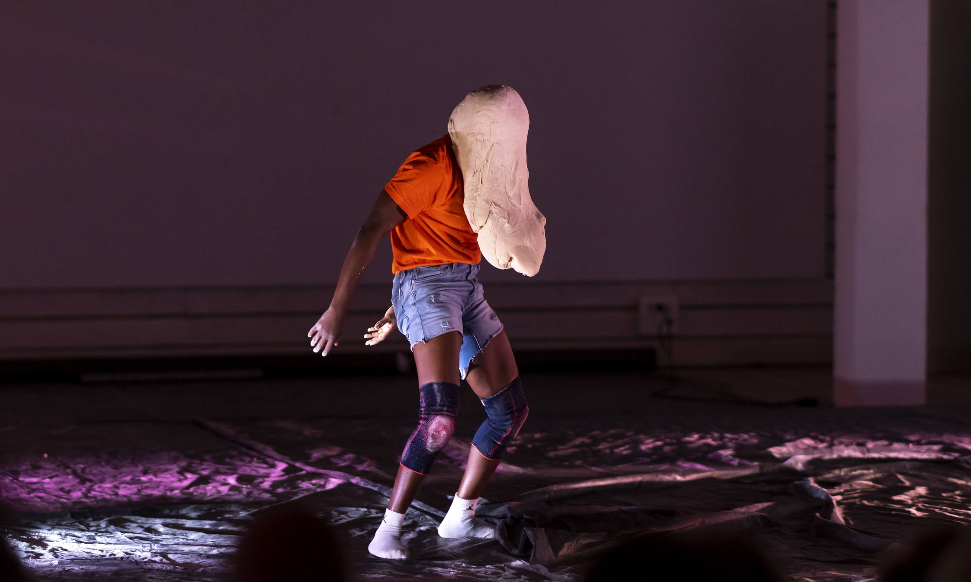 Eine Person in einem orangenen T-Shirt, einer Jeansshorts und Knieschonern gekleidet tanzt auf einer mit schwarzer Plastikplane verdeckten Bühne. Das Gesicht der Person ist von einer Teigmasse bedeckt.