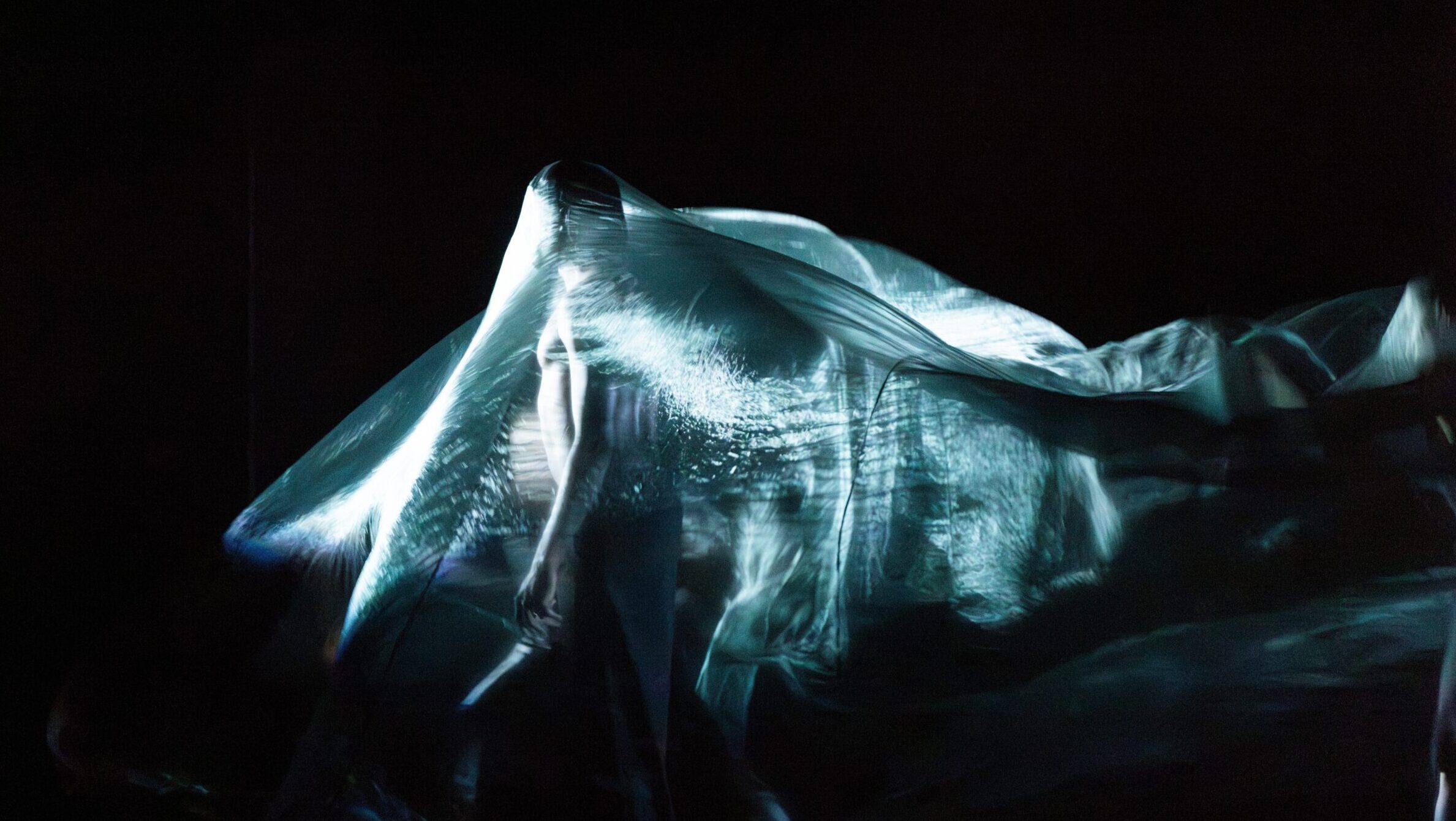 Person steht unter einem großen, transparenten, silberblau schimmerndem Tuch auf einer dunklen Bühne während einer Theaterperformance der Compagnie 111.